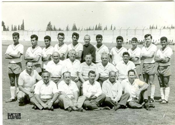 1961/62 - גביע - סיבוב 5 - הפועל כפר בלום (2:10) - תמונה קבוצתית שצולמה לפני המשחק מול הפועל כפר-בלום בגביע המדינה. עומדים, מימין: אבשלום רצאבי, נחום סטלמך, בועז קופמן, אלקנה שמן, זכריה רצאבי, יעקב ויסוקר, שלמה נהרי, אריה רדלר, דוד אטואר, אמנון מזרחי, שלמה נומה. באמצע יושבים מימין שבתאי זילברשטיין וחברי ההנהלה שלום קולקר, מנחם הלר, מאיזנר, אביגדור. למטה חברי אגודת הסדרן ומשמאל כורע המעסה מאיה.